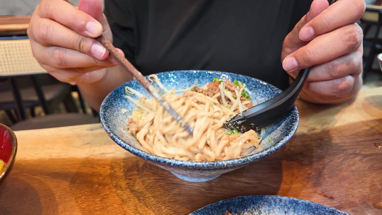 Primo piano di mani che mescolano noodles con salsa al sesamo e carne di maiale macinata in una ciotola blu