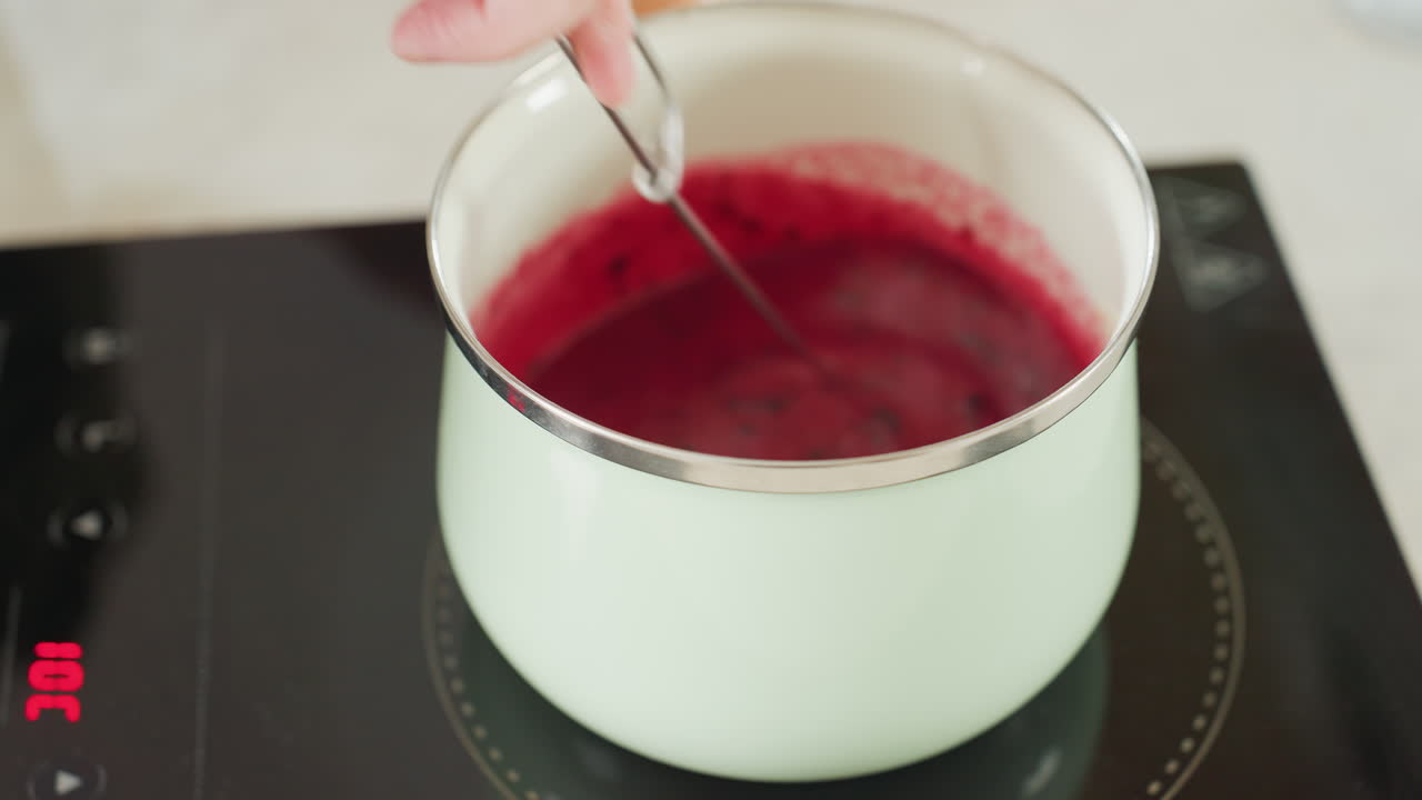 Hand holding metal twister stirs red soup inside pastel green pot on electric stove with digital heat display, showing cooking process in clean modern kitchen
