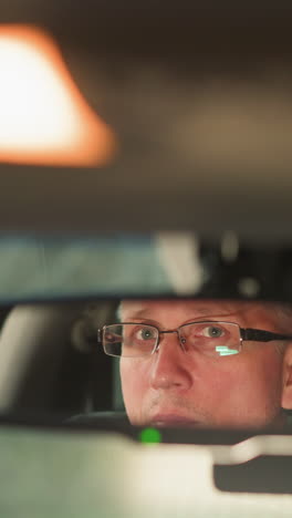a man wearing glasses is reflected in the rearview mirror of a car. his expression is focused and contemplative, as the soft interior lighting illuminates the scene