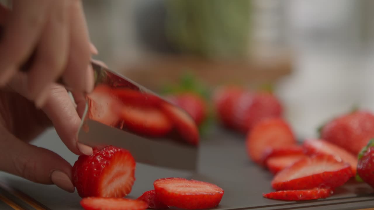 Uma mulher usando esmalte pêssego claro pica lentamente um morango vermelho e maduro em uma tábua de corte