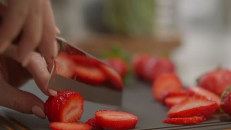 a woman wearing light peach nail polish slowly chops up a red, ripe strawberry on a cutting board