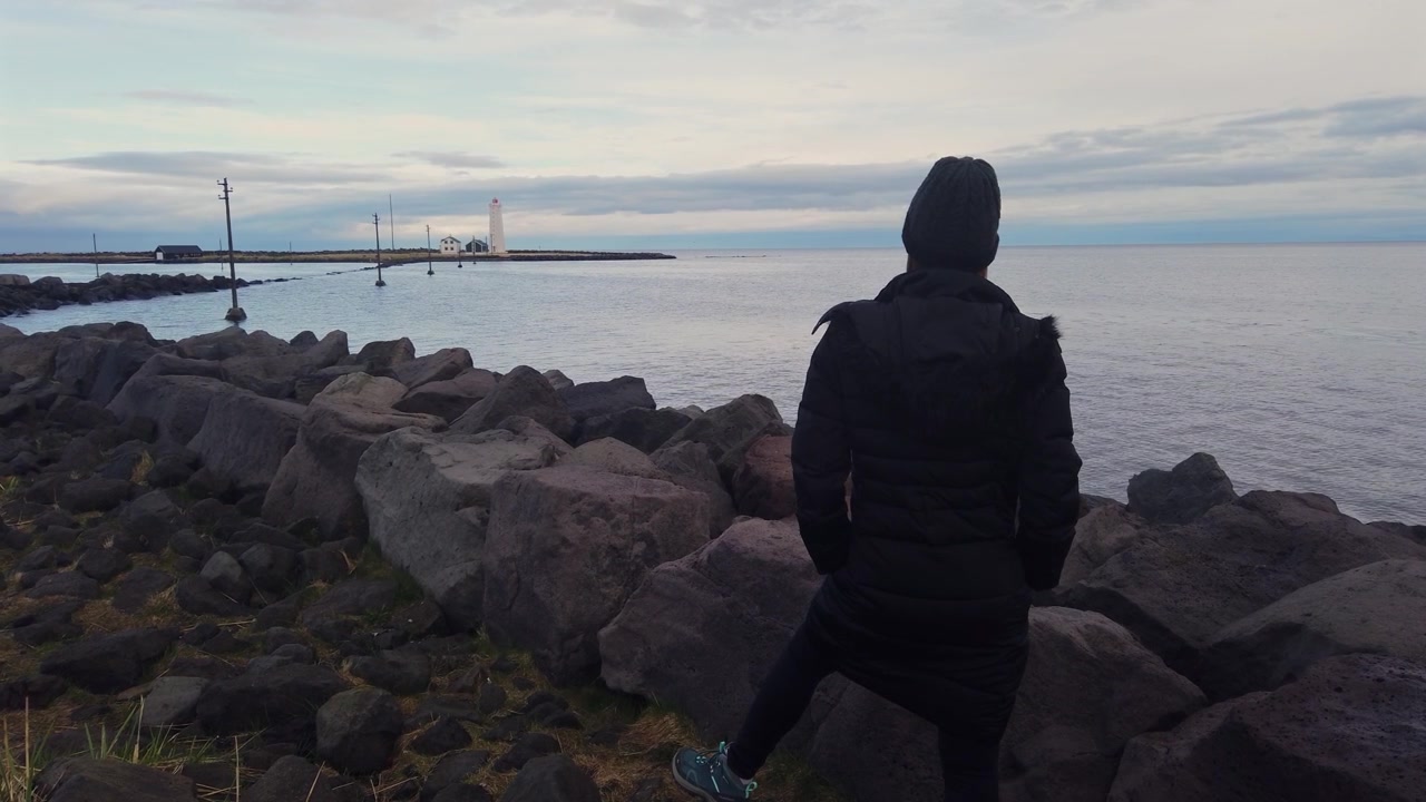 Vista posteriore di una donna solitaria in piedi su una diga frangiflutti rocciosa, che osserva pensierosa il mare e il lontano faro di Akranes in Islanda.