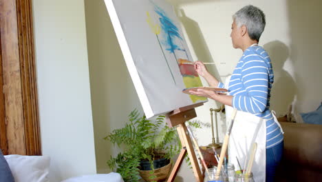 Mujer-Birracial-Senior-Enfocada-Usando-Delantal-Y-Pintando-Sobre-Lienzo-Grande-En-Casa,-Cámara-Lenta