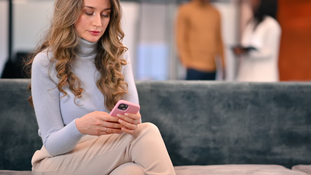 En kaukasisk kvinne bruker smarttelefonen sin mens hun sitter på en sofa på et kontor
