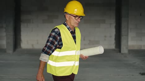 Caucasian-senior-engineer-walking-and-holding-plans-on-construction-site.