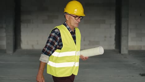 caucasian senior engineer walking and holding plans on construction site.