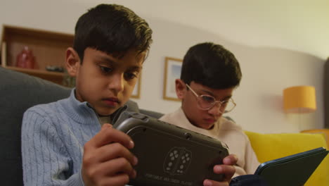 two young boys sitting on sofa at home playing games or streaming onto digital tablet and handheld gaming device 3