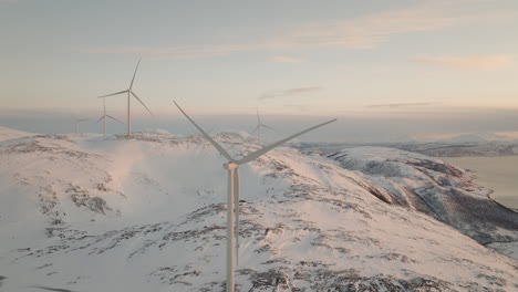 Vista-Aérea-De-La-Montaña-Nevada-De-Las-Palas-De-La-Turbina-Eólica-Girando,-Puesta-De-Sol-ártica