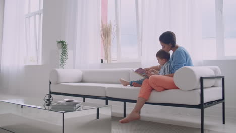 a young mother with a child reading a book sitting in a bright white interior of the house in the living room on the couch