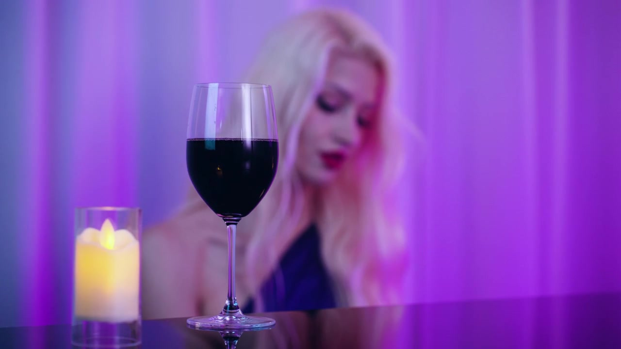 Bicchiere di vino rosso accanto a una candela con una bella donna bionda in abito che suona il pianoforte in un pub sfocato sullo sfondo