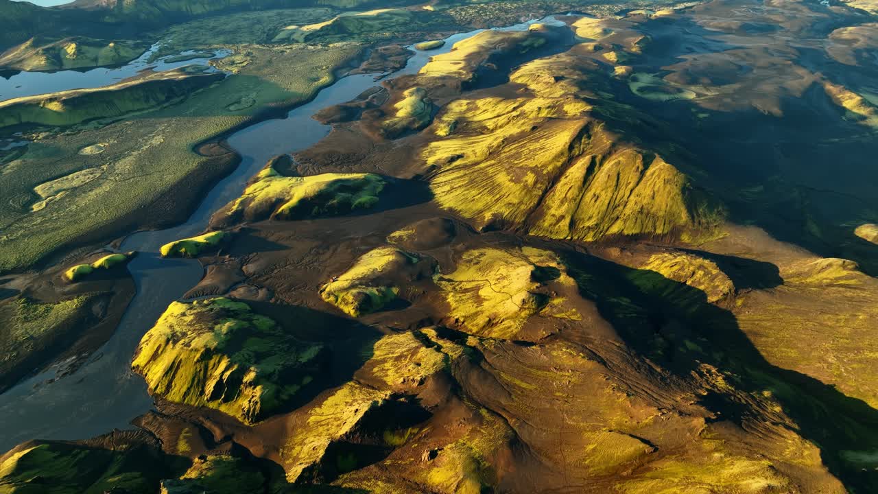 Vista aérea de un dron sobre una cresta en las tierras altas de Islandia con colinas volcánicas cubiertas de musgo y un río serpenteante, luz cálida de la hora dorada y largas sombras que dan forma a un paisaje remoto épico.