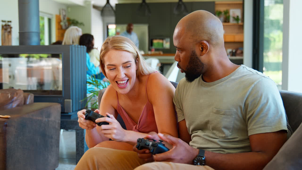 가정에서 소파에 앉아 컴퓨터 게임을 하는 부부, 배경에는 여러 세대의 가족이 있습니다.