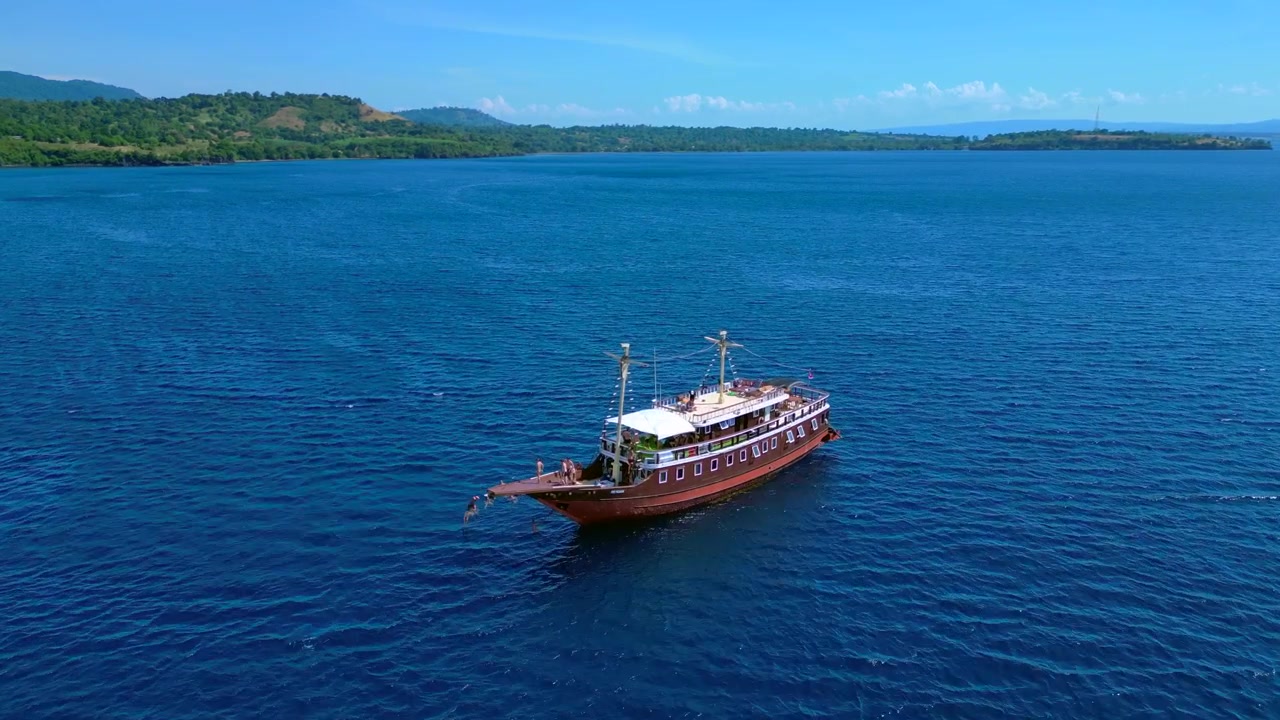 Perahu pinisi tradisional di perairan terbuka dengan seseorang melompat dari haluan ke laut. Pemandangan udara menawan, panorama drone yang terbang maju.