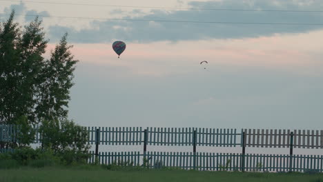 distant view of paraglider and hot air balloon gliding past power lines and tree branches above rural road fence under pink dusk sky conveying serene adventure mood over open landscape