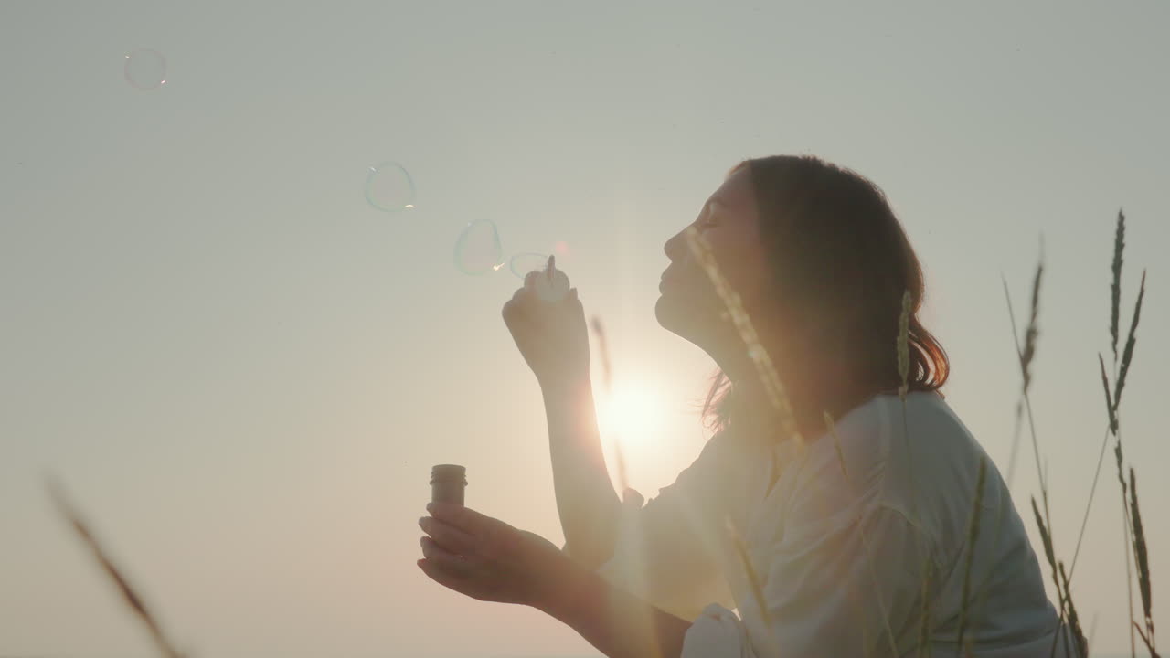 Lady gently blowing bubbles in grassy field as golden sun glows behind her silhouette, surrounded by tall grain stalks in peaceful atmosphere