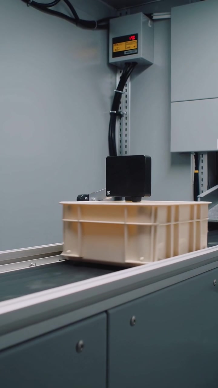 Vertical video: Moving beige crate along conveyor under black sensor in bay signaling control box