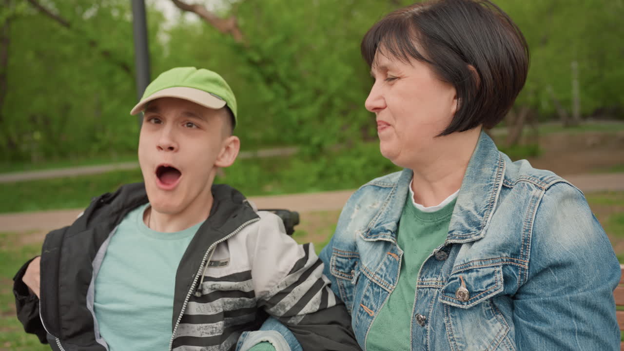 Young Man In Wheelchair And Caregiver Share Joyful Laughter On Bench In Park, Open Smiles And Playful Gestures, Sunlight Through Trees, Relaxed Upbeat Mood, Authentic Companionship