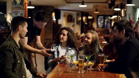 cheerful students are paying with smartphone while sitting at table in nice cafe. quick easy contactless payment concept. attractive people and modern bar interior are visible.