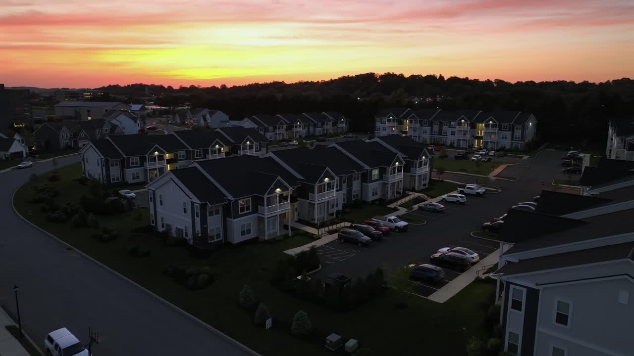 Aerial view of American suburban neighborhood at sunset. Rows of modern apartment buildings with gabled roofs surround parking areas. The vibrant sky adds warmth, creating a peaceful evening scene.
