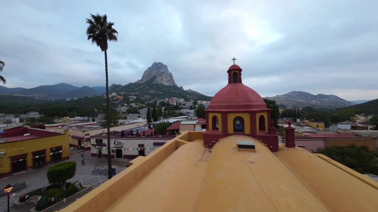 Piazza storica con il monolito Peña de Bernal sullo sfondo, Queretaro all'alba, sorvolo dinamico con drone su edifici gialli e rossi