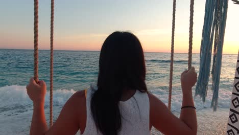 back of young and carefree girl in summer dress swinging in front of ocean