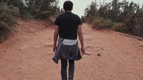 Young-man-walks-down-dusty-dirt-road-in-desert-on-hike,-slow-motion