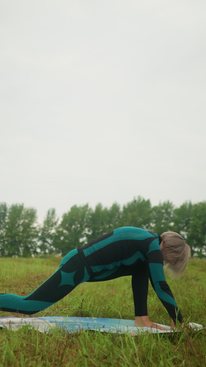 vista lateral de una mujer en traje verde y negro al aire libre en un vasto campo de hierba en una alfombra de yoga practicando una postura baja, bajo un cielo nublado con árboles alineados en el fondo