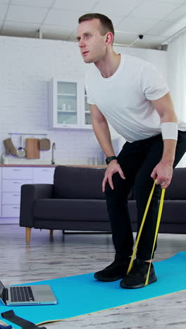 concentrated man exercising with a rubber band at home. young man doing working out while standing on a mat in the kitchen. sportive lifestyle during quarantine. vertical video