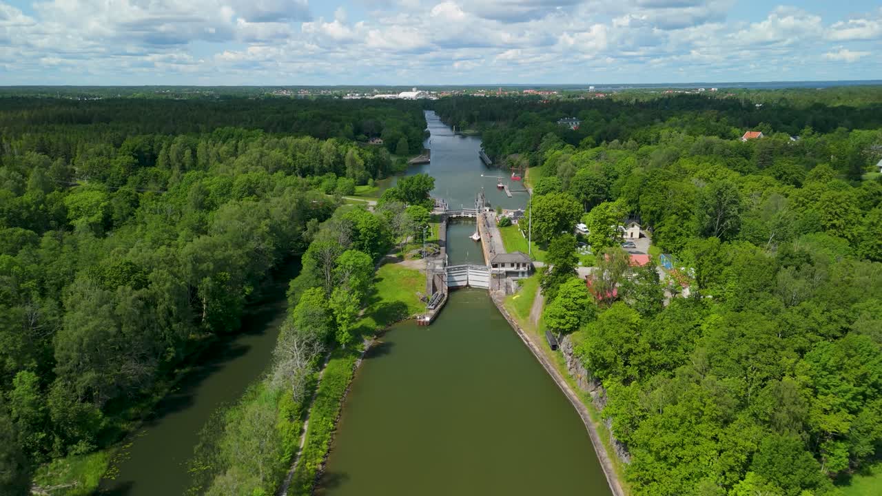 Sluizen van Vänersborg, Zweden — luchtopname van historische kanaalsluisdeuren die openen op het Götakanaal, met passerende boten, weelderige bosoevers, een sluiswachtershuis en een stad zichtbaar aan de horizon in de zomer.