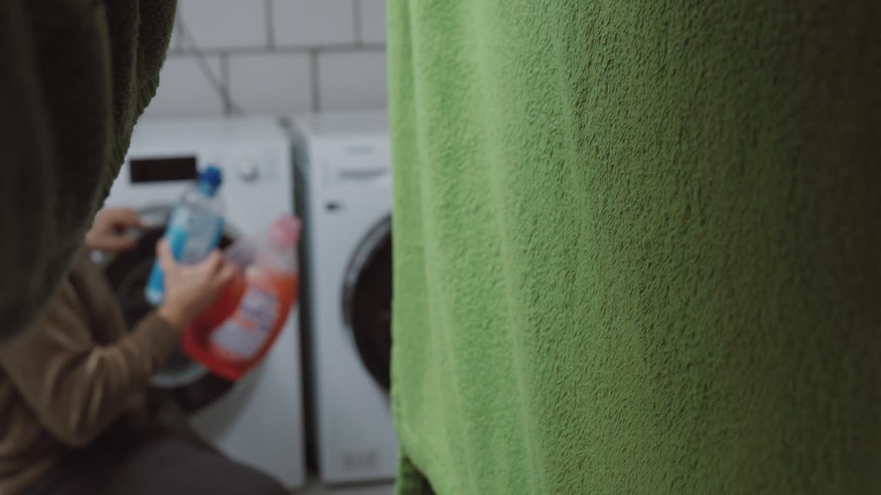 Muž klečí před pračkou a drží prací prostředek, nejistý, který použít. Koncept každodenního rozhodování, domácích prací, zmatku a domácí rutiny