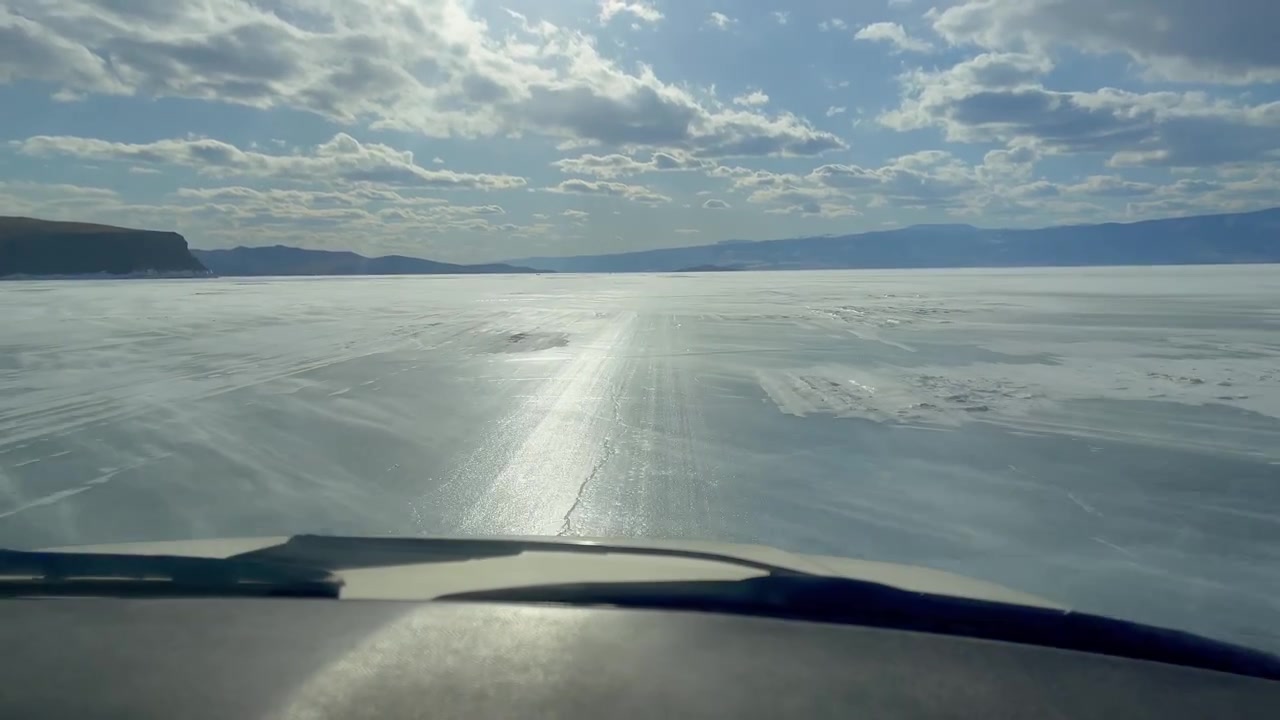 Vista POV da un veicolo che guida sulla superficie ghiacciata del Lago Baikal con il riverbero del sole, ghiaccio strutturato e montagne distanti sotto un cielo nuvoloso in Russia