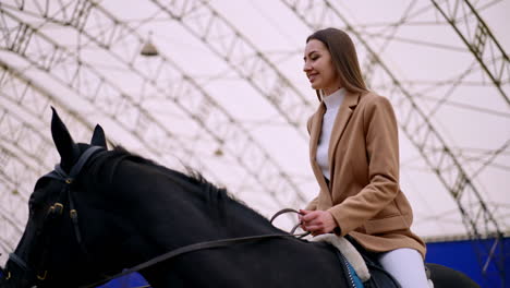 positive smiling caucasian lady in beige jacket riding a black horse. low angle perspective. blurred drill-hall at backdrop.