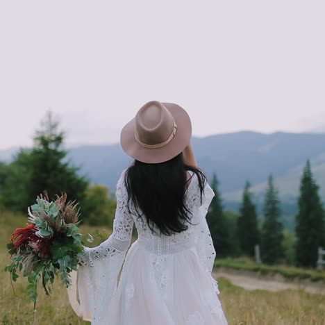 Backside view of a woman in wedding dress among nature. Bride in white dress with sleeves and a hat on the background of mountains.