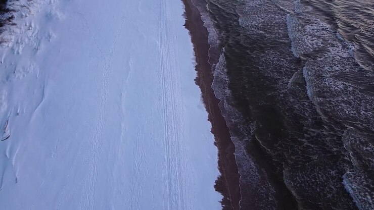 Beautiful romantic aerial revealing shot flying over a snow covered beach after the sunset on the calm Baltic sea coastline, high contrast sky, wide angle drone shot moving forward, tilt up