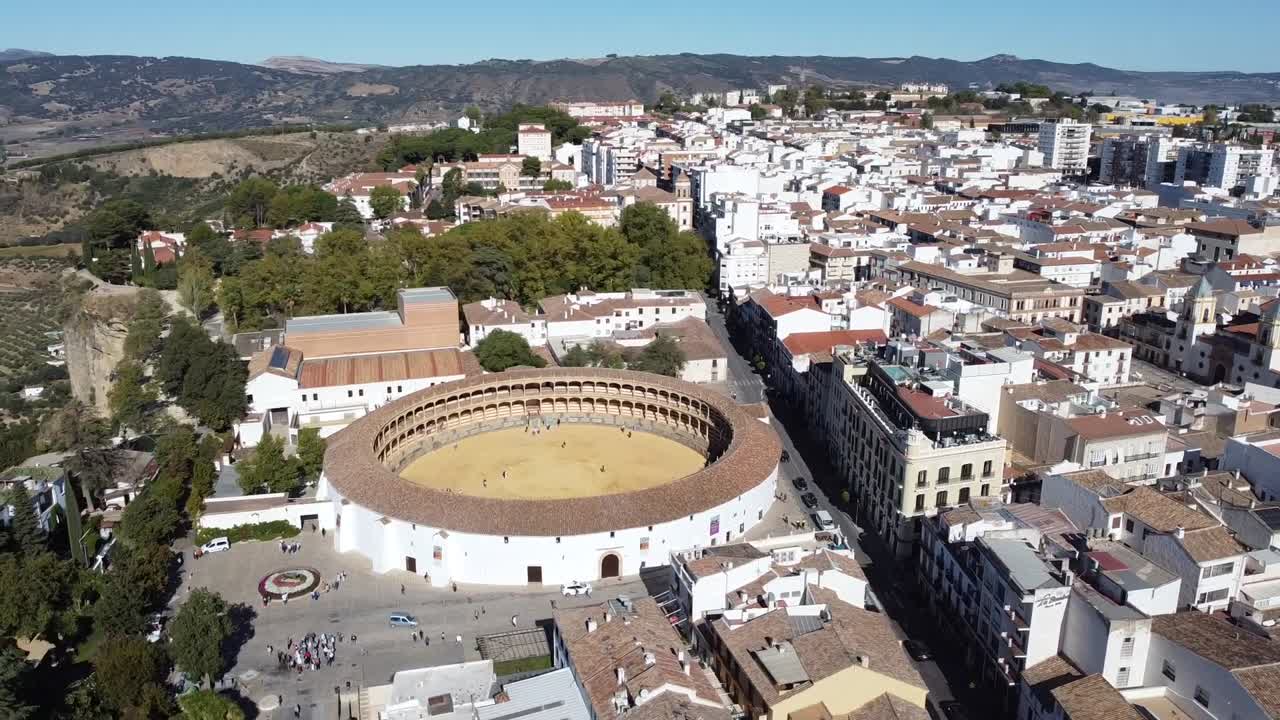 Drone shot of circular bullring in center of town with surrounding white buildings, narrow streets, and distant hills, capturing contrast between architecture and natural terrain