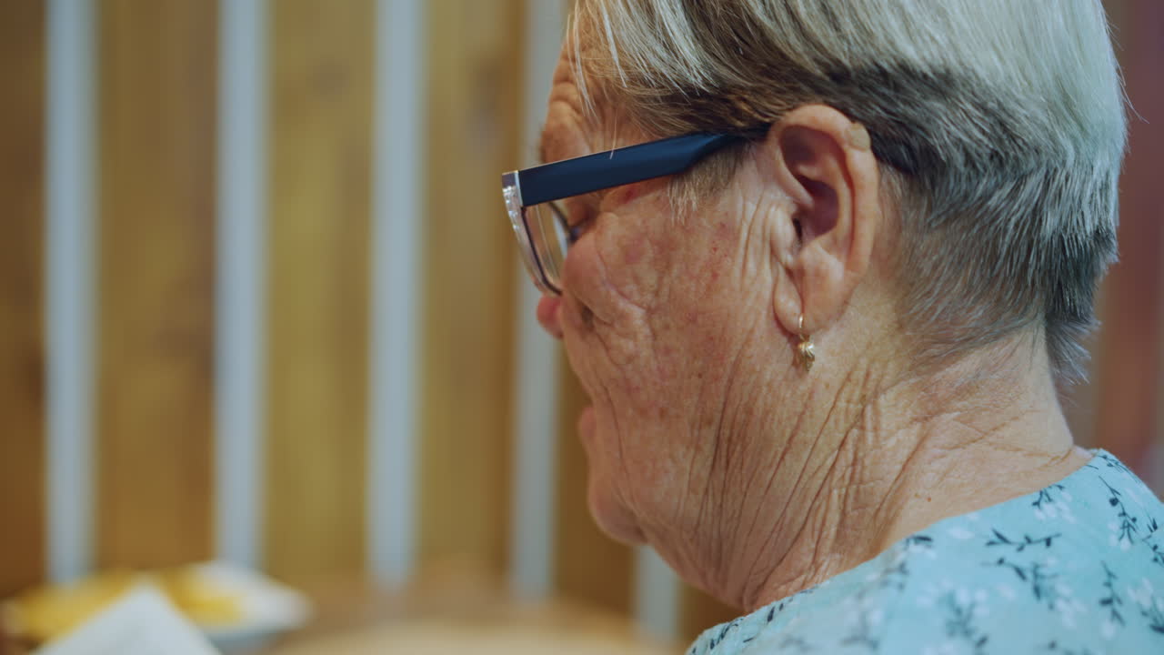 Nonna bianca di profilo dall'aria pensierosa, sfondo con pannelli in legno, occhiali da lettura, piccolo orecchino visibile, calda vignetta da tavolo con piatto, espressione contemplativa, luce interna soffusa, serena.