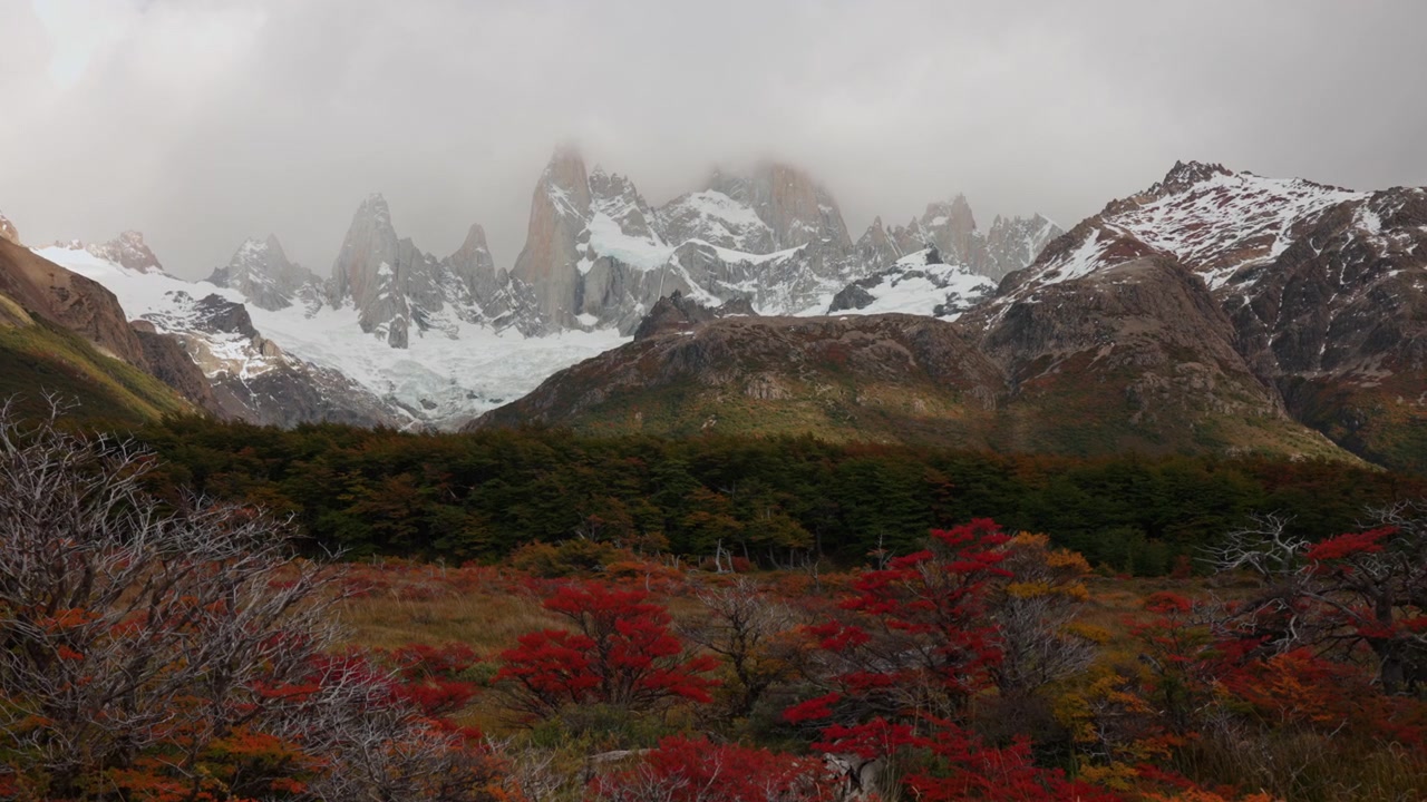 Una vista di montagne frastagliate coperte di nuvole, circondate da alberi autunnali colorati nei toni del rosso e dell'arancione nella regione della Patagonia