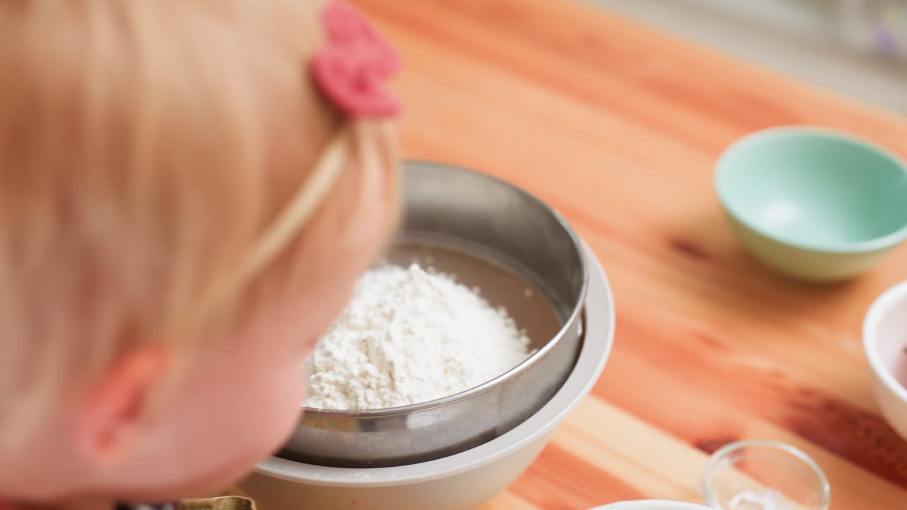 Close up of little girl in checkered dress with hairband pouring flour from small bowl into metal sift, concentrating carefully while engaging in early cooking practice on wooden table