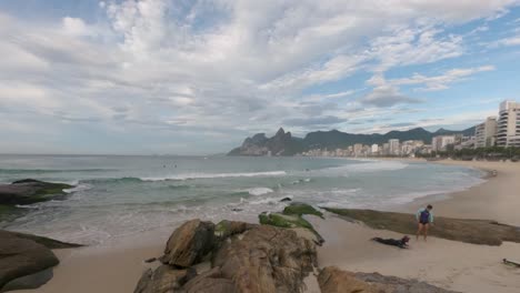 time lapse early morning ipanema beach in rio de janeiro with rocks scattered in the foreground and the two brothers mountain in the background against a blue cloudy sky with sunlight coming in