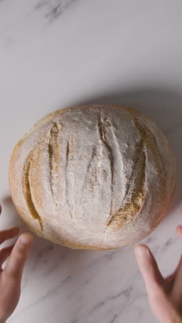 Vertical-Video-Overhead-Shot-Of-Freshly-Baked-Loaf-Of-Bread-Being-Put-Down-Onto-Marble-Work-Surface