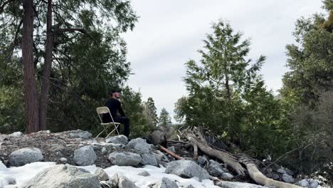 a young caucasian male dressed in black hiking clothes and a black ball cap walking across snow and rocks in the forest to sit on a plastic chair and contemplate life on an overcast day