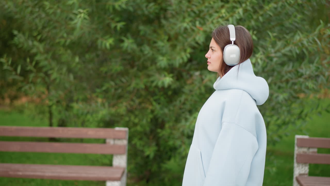 Pretty woman in casual wear strolling through garden wearing white headphones, with background featuring flowers and concrete bench, ideal for outdoor walks, nature, or relaxation scenes