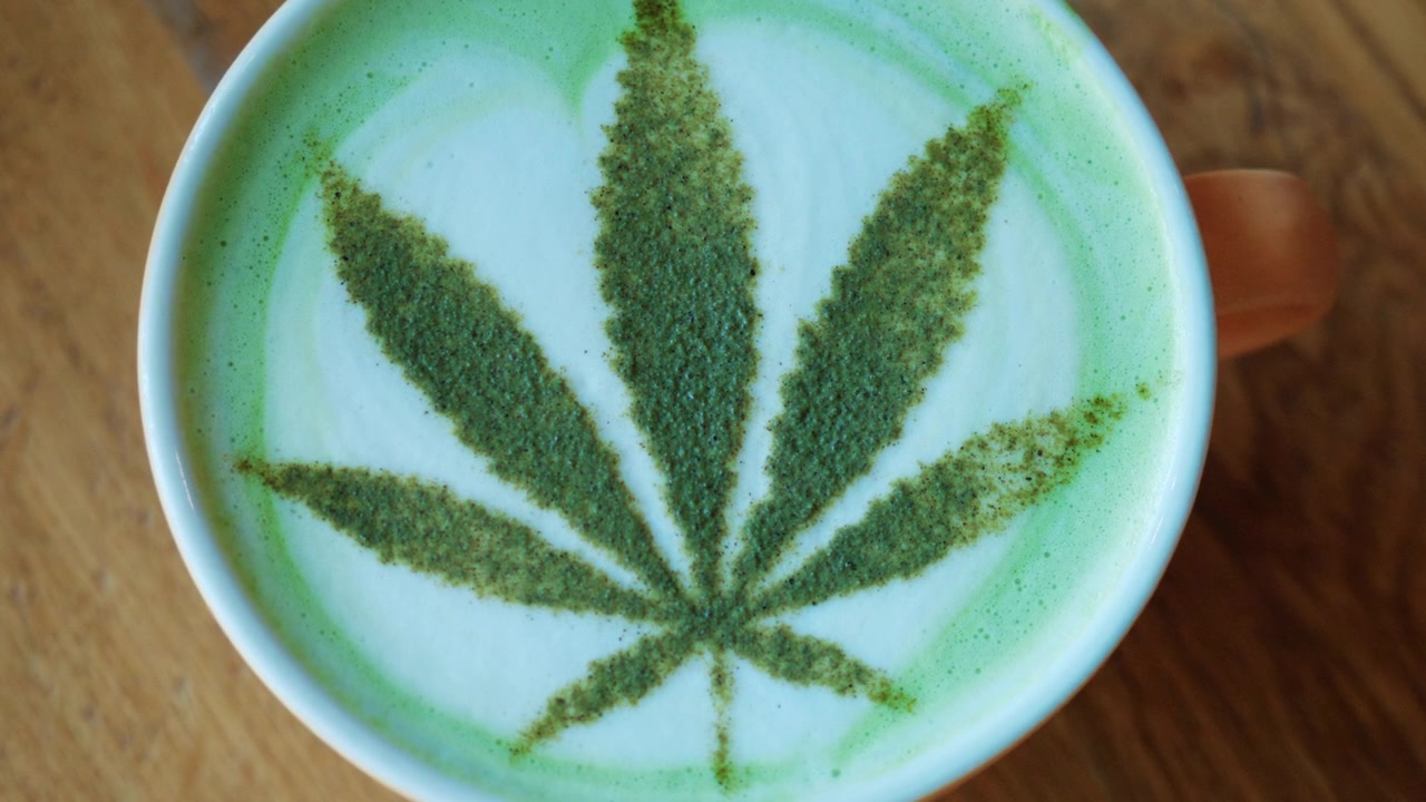 Vista dall'alto di un matcha latte in una tazza di ceramica, decorato con tè verde in polvere che forma un motivo a foglia che ricorda una foglia di cannabis.