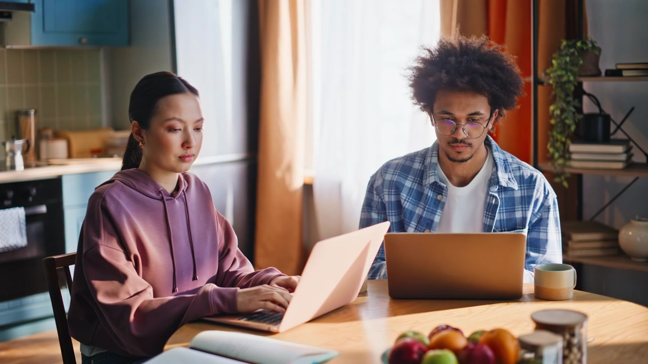 Multiethnic couple working laptops at home kitchen table. Remote freelancers.
