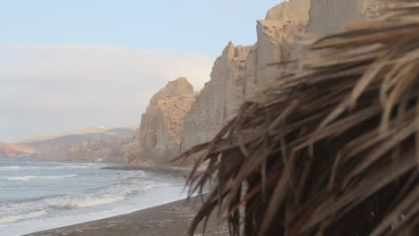 detalle de una sombrilla de playa de paja con la playa negra y formaciones de acantilados blancos en el fondo