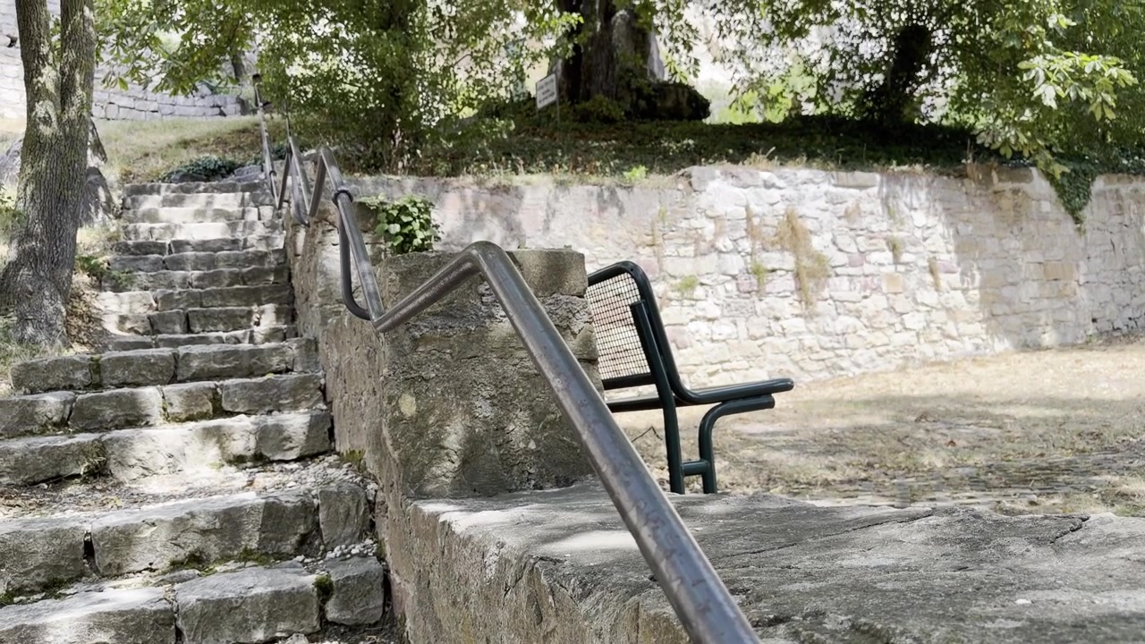 Uma inclinação lenta para cima revela uma escadaria de pedra histórica com luz solar difusa, corrimão de ferro forjado e um banco que leva ao famoso Isteiner Klotz. Plano de Tilt para Cima