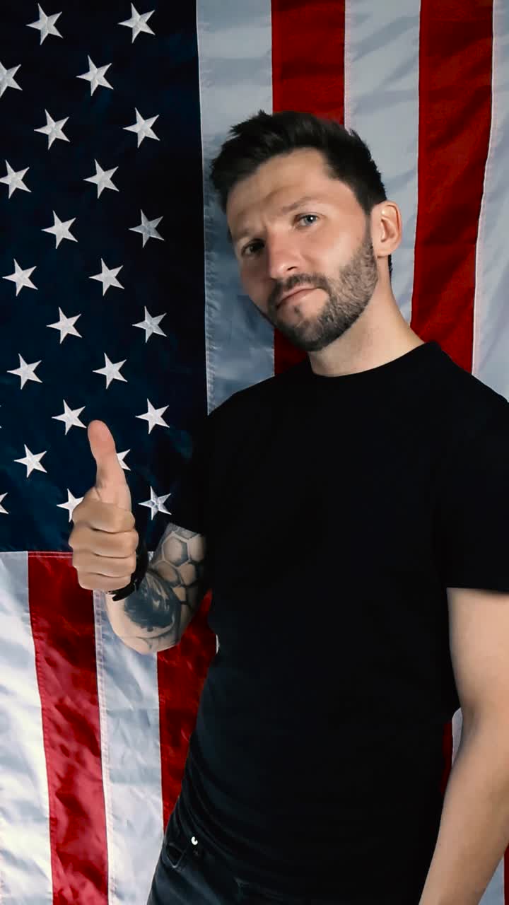 Tattooed patriot doing thumbs up expression in front of vertical United States of America flag with happy and approving emotion while nodding into the camera. Man has short beard and is white