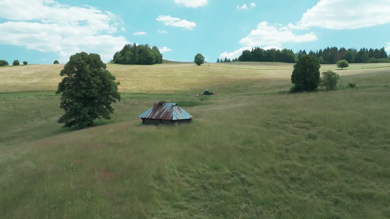 드론이 떠다니며 경치 좋은 시골 풍경을 촬영합니다. 언덕이 있는 지형, 높은 흔들리는 잔디, 그리고  ⁇ 은 지붕을 가진 시골의 창고