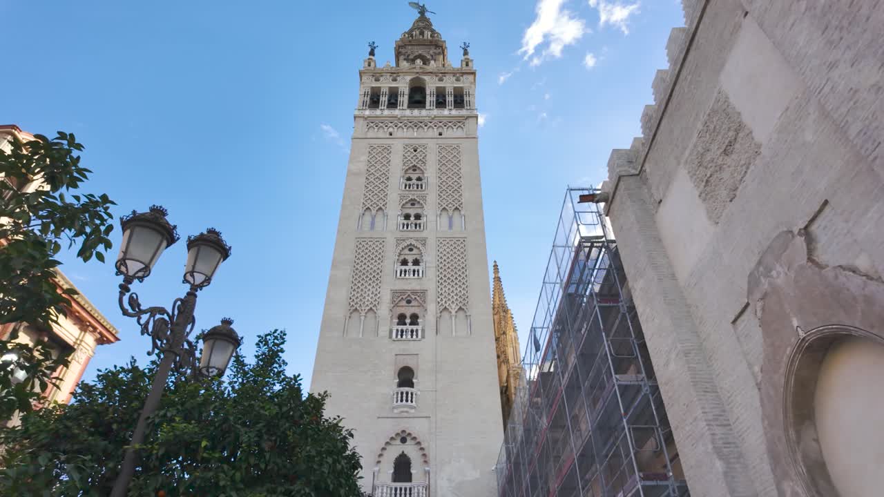 Giralda, the imposing bell tower of Seville Cathedral, stands tall against a vibrant blue sky, showcasing its intricate Moorish architecture and rich history. POV push forward shot
