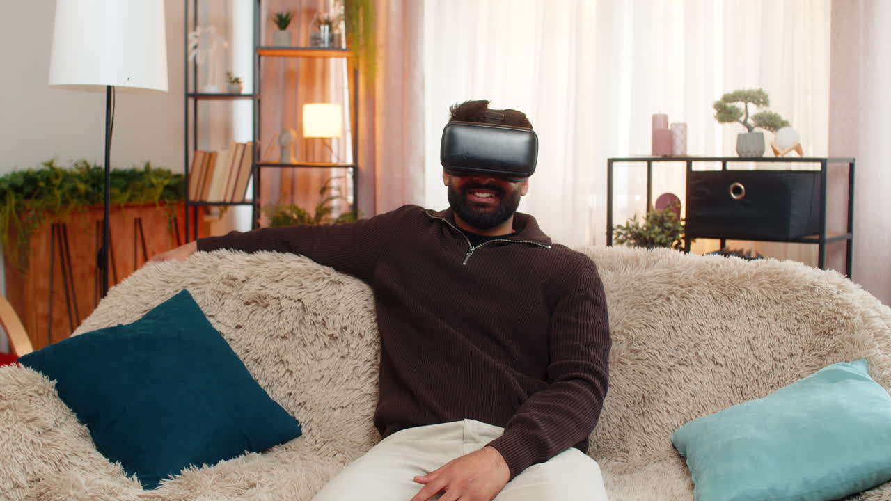 Indian man on sofa wears vr headset at home and reacts with excitement to immersive virtual world